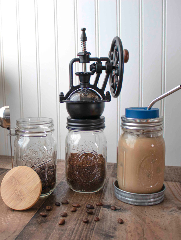lifestyle photo of coffee spoon clip, bamboo stopper lid, vintage reproduction coffee grinder, deep blue silicone straw hole lid, and a medium thin bent stainless steel straw on regular mouth pint mason jars