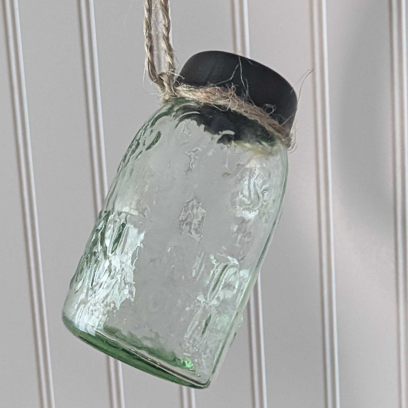 mini green glass jar ornament with a black lid hanging against a white paneled wall.