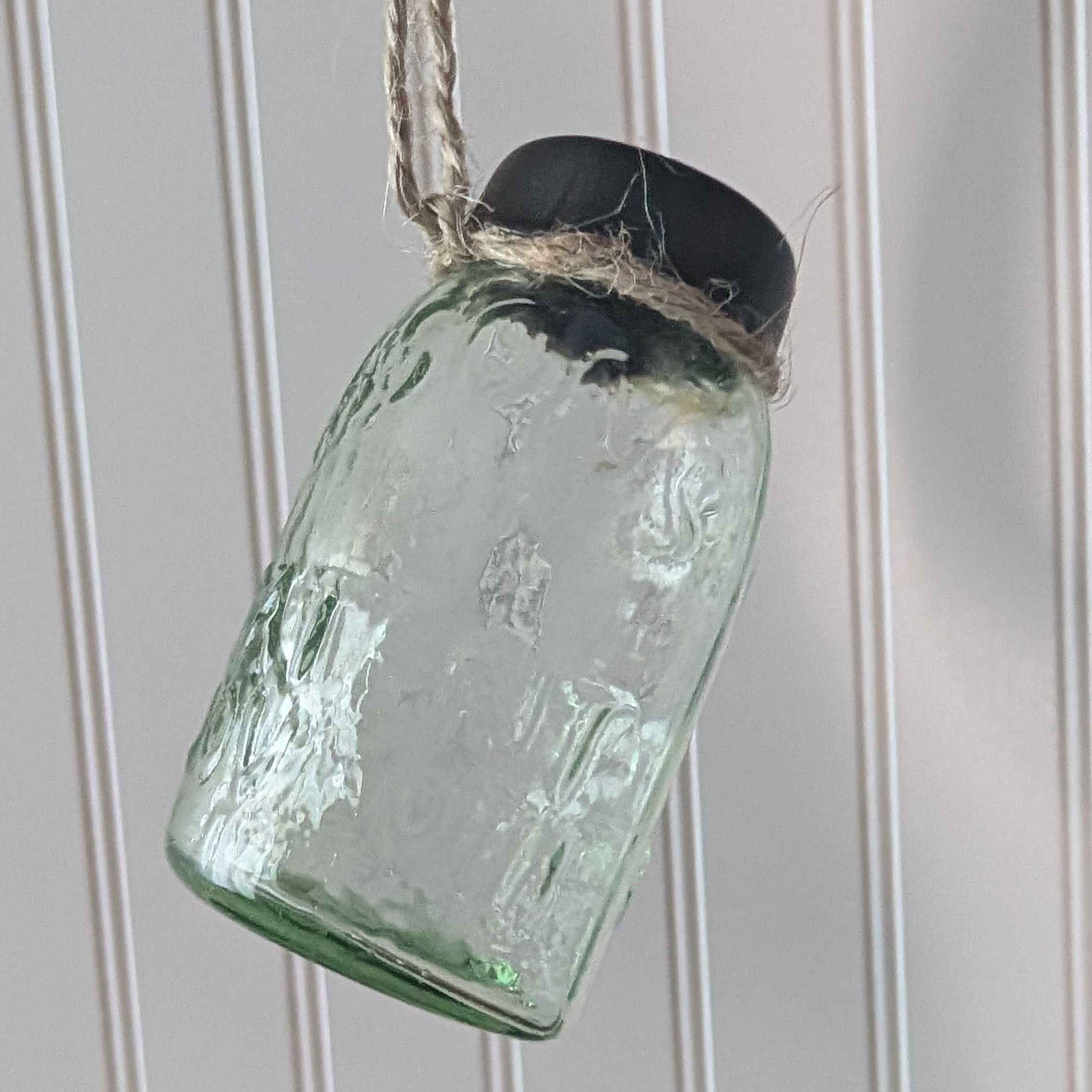 mini green glass jar ornament with a black lid hanging against a white paneled wall.