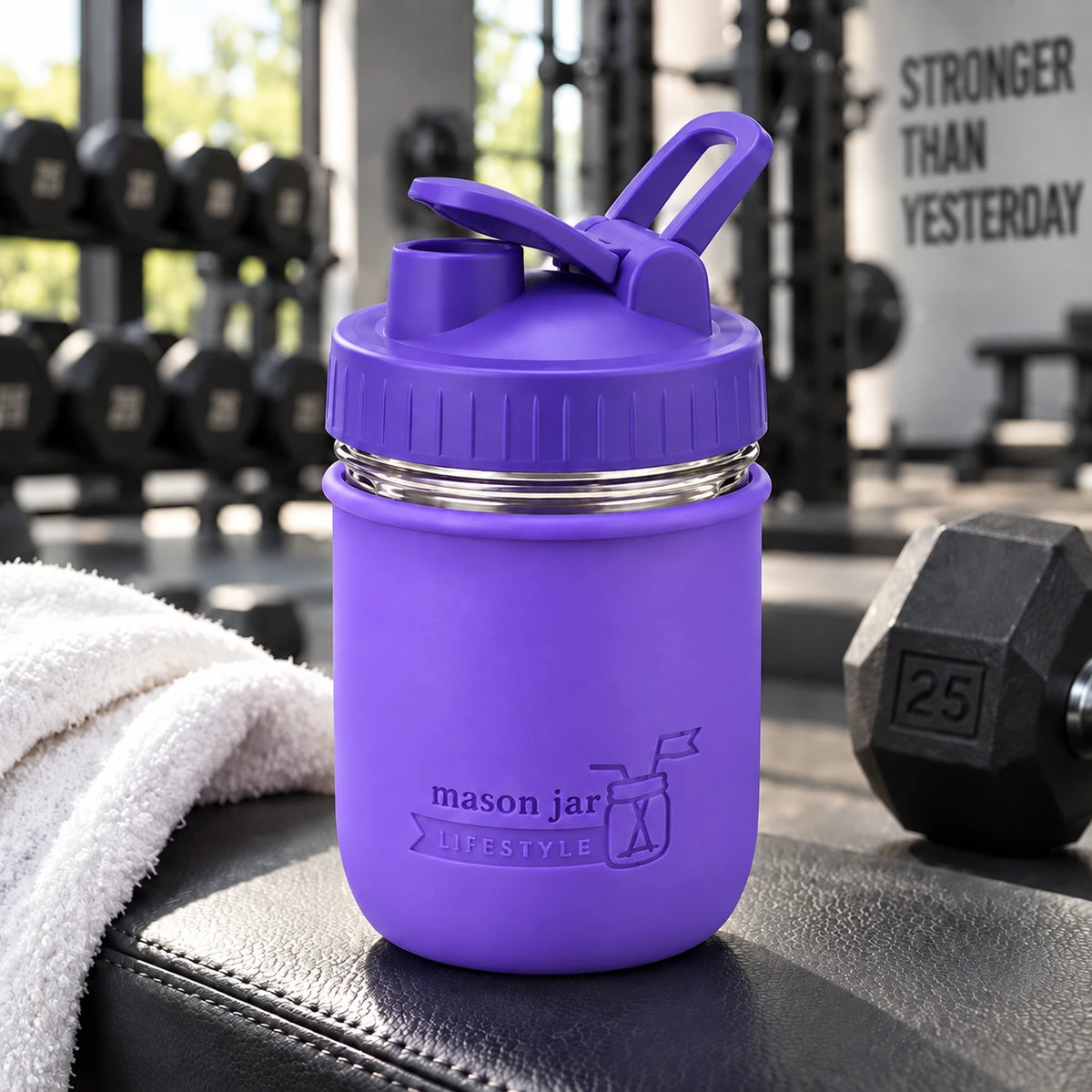 Mason Jar Lifestyle 16oz glass protein shaker bottle with Ultra Violet silicone sleeve and carry-loop lid on a gym bench beside a towel and dumbbell.