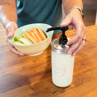 A woman pressing down on a food grade pump with ranch into a bowl of carrot sticks and cucumbers.