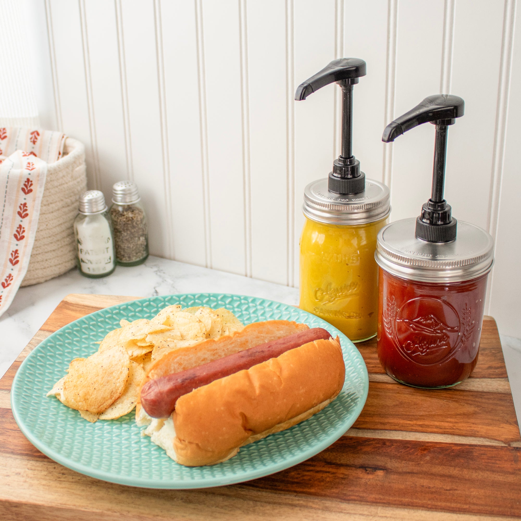 A staged photo of the food pump on regular mouth and wide mouth Mason jars, next to a plate with a hotdog and chips.