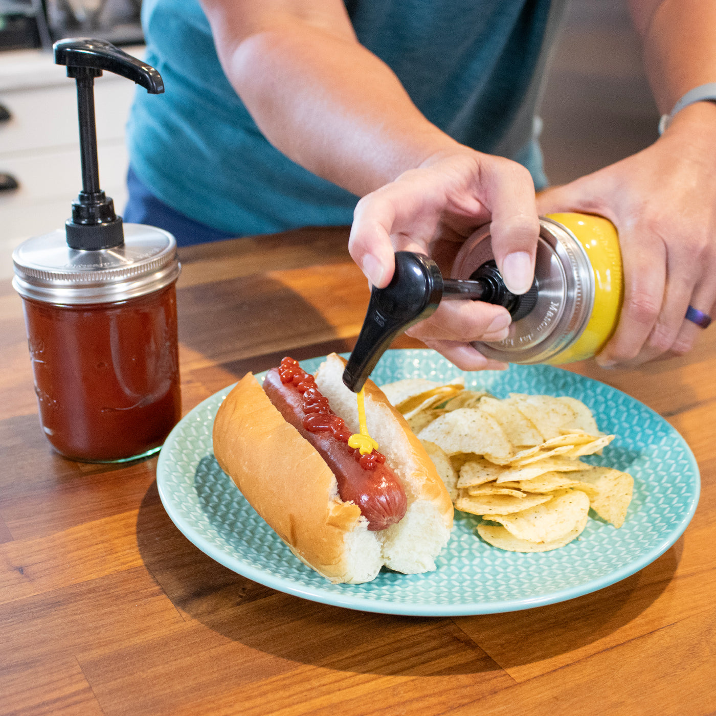 A person  pressing down on the food grade pump over a hotdog, dispensing mustard.