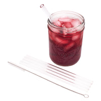 4 pack of medium glass straws and the straw cleaner next to it, laying in front of a Mason jar full of juice, with a glass straw in it.