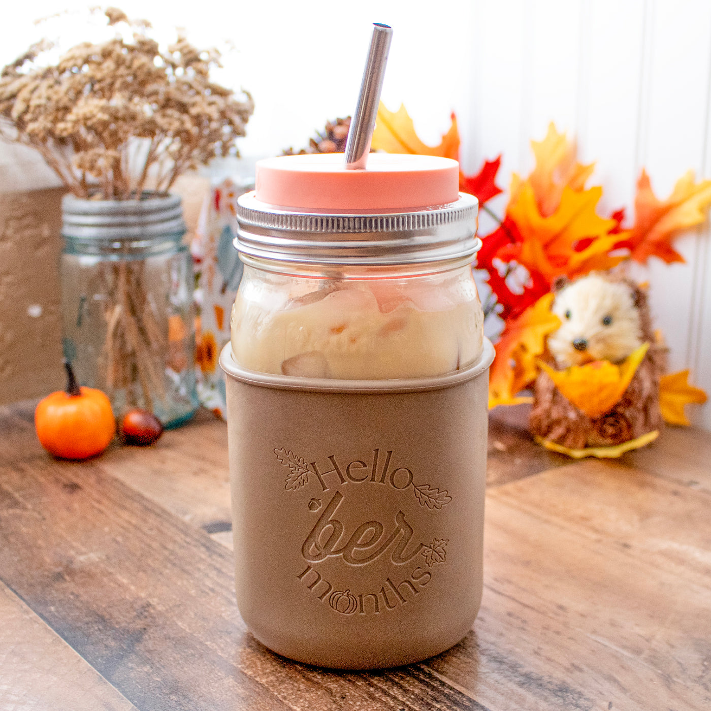 A Mason jar quart jar with the Hello Ber Month sleeve and coral pink straw hole lid, in front of a fall decor.