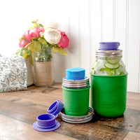 Two regular mouth Mason jars with silicone chug lids on them, in bright blue and ultra violet. An open one lays to the side of them, showing off the small silicone strap that connects the two sides, so you don't have to worry about losing the top.