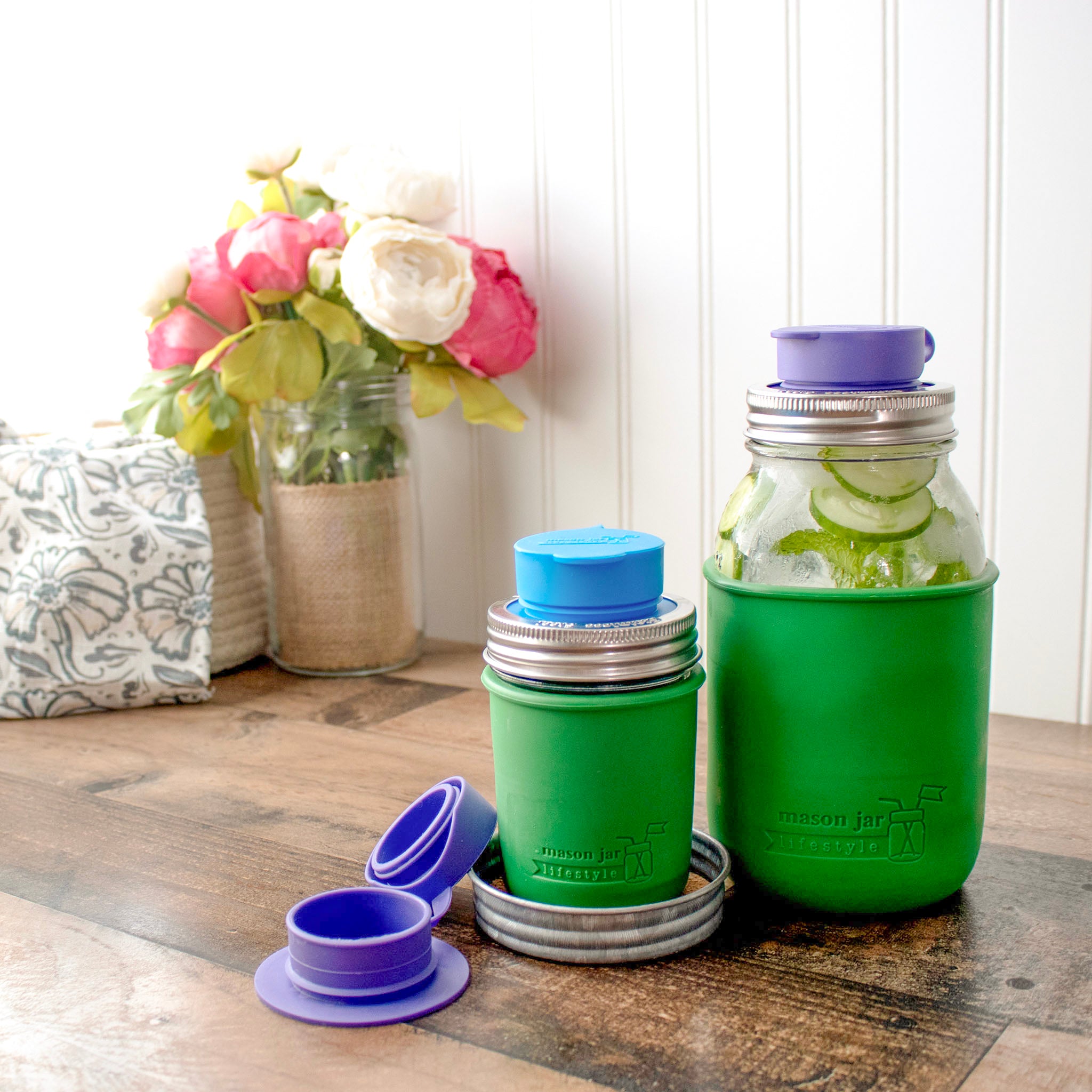 Two regular mouth Mason jars with silicone chug lids on them, in bright blue and ultra violet. An open one lays to the side of them, showing off the small silicone strap that connects the two sides, so you don't have to worry about losing the top.