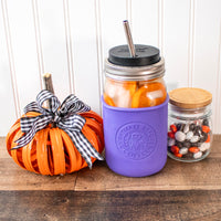The laser design on a quart Mason jar ultra violet colored sleeve. The jar is surrounded by cute Halloween decor:  a metal pumpkin made up of Mason jar rings, and a small jar of colorful chocolate candies.