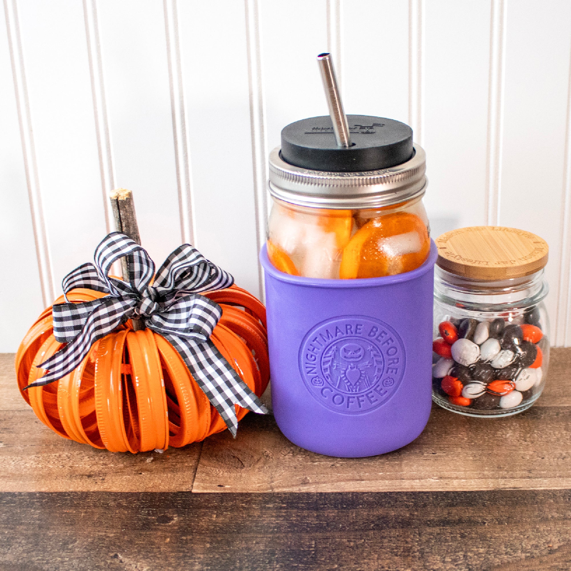 The laser design on a quart Mason jar ultra violet colored sleeve. The jar is surrounded by cute Halloween decor:  a metal pumpkin made up of Mason jar rings, and a small jar of colorful chocolate candies.