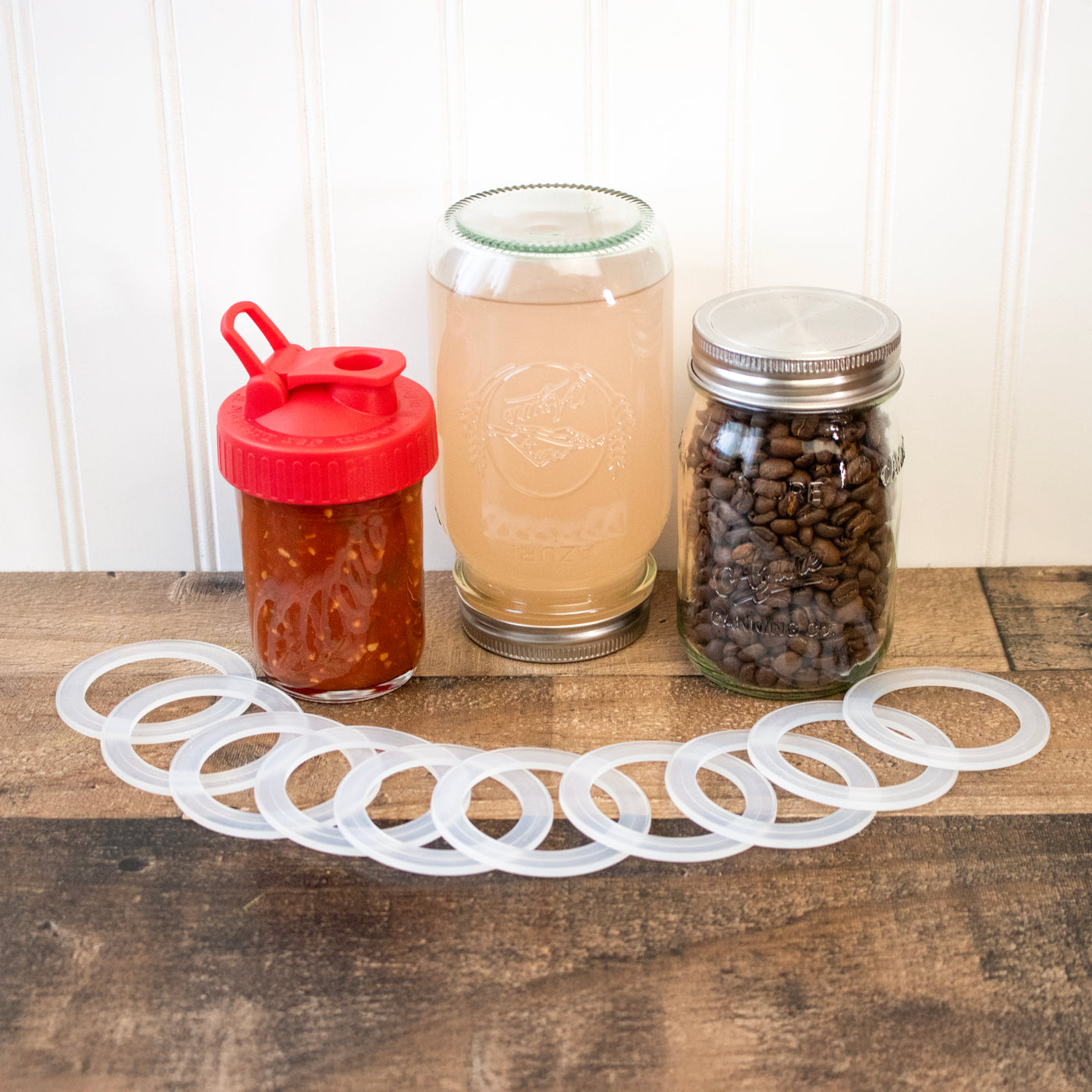 Regular mouth seals laid out on a darker wood surface so you can see the ridges that help keep the seal in place. Behind are three jars with lids with silicone rings in them, one is upside down to show the sealing power.
