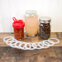 Regular mouth seals laid out on a darker wood surface so you can see the ridges that help keep the seal in place. Behind are three jars with lids with silicone rings in them, one is upside down to show the sealing power.