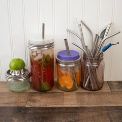 Two different wide mouth Mason jars with silicone straw hole lids on them, with reusable straws. a juicer and jar of straws sit next to them.