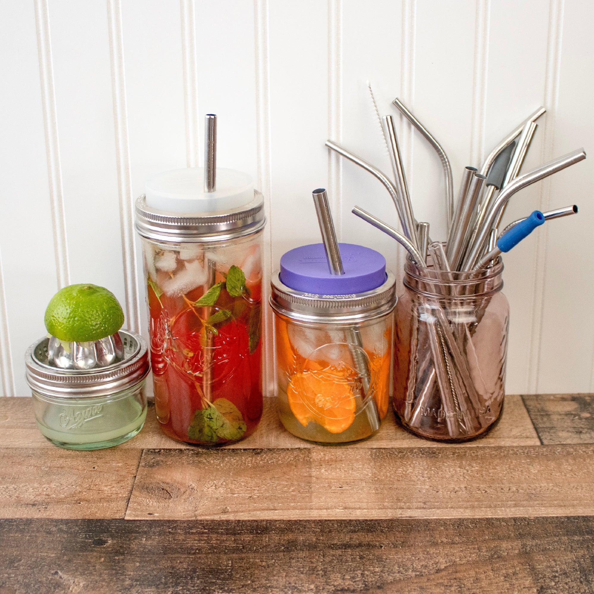 Two different wide mouth Mason jars with silicone straw hole lids on them, with reusable straws. a juicer and jar of straws sit next to them.