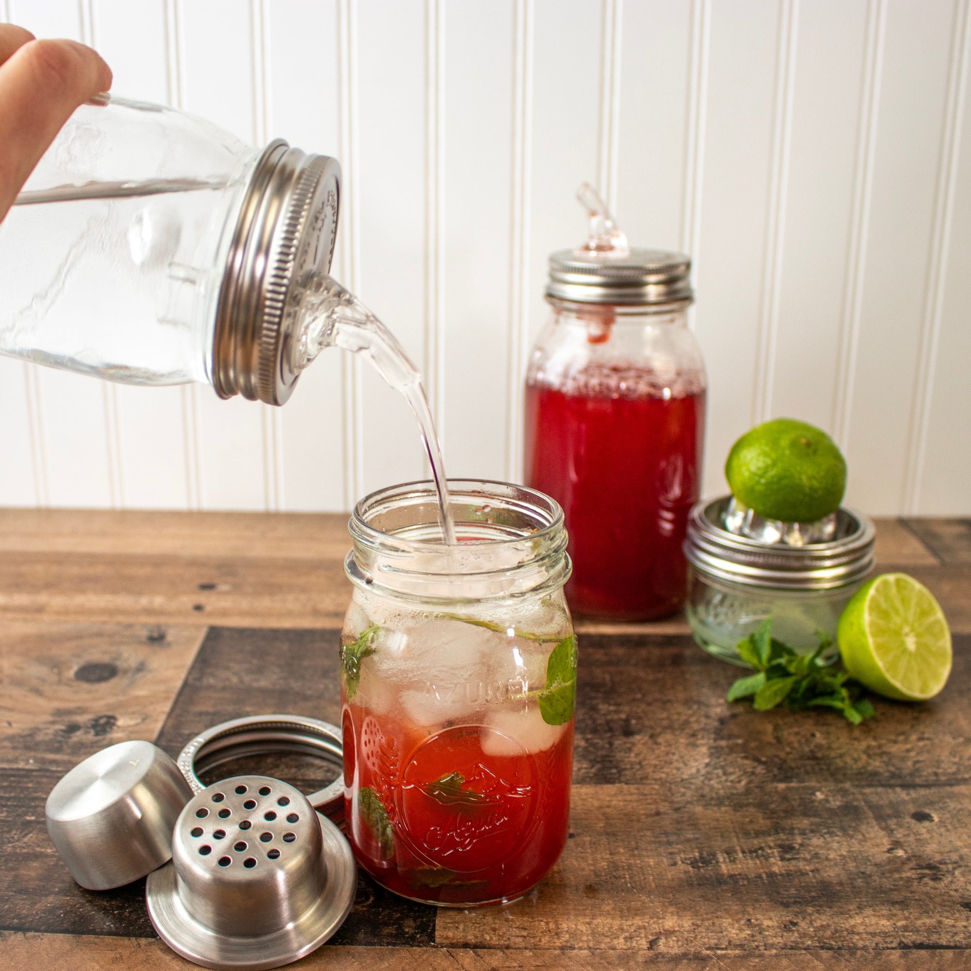 Pieces of the cocktail shaker, next to a drink, with strawberry syrup being poured into it.