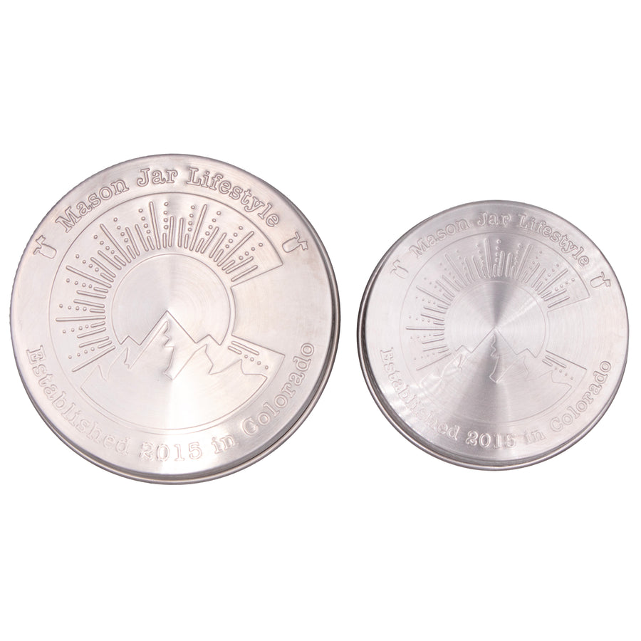 Two stainless steel storage lids for Mason jars up close, showing off the design that says "Mason Jar Lifestyle" and "Established 2015 in Colorado" with a big "C" in the middle with a silhouette of a mountain, a sun, and sun rays.