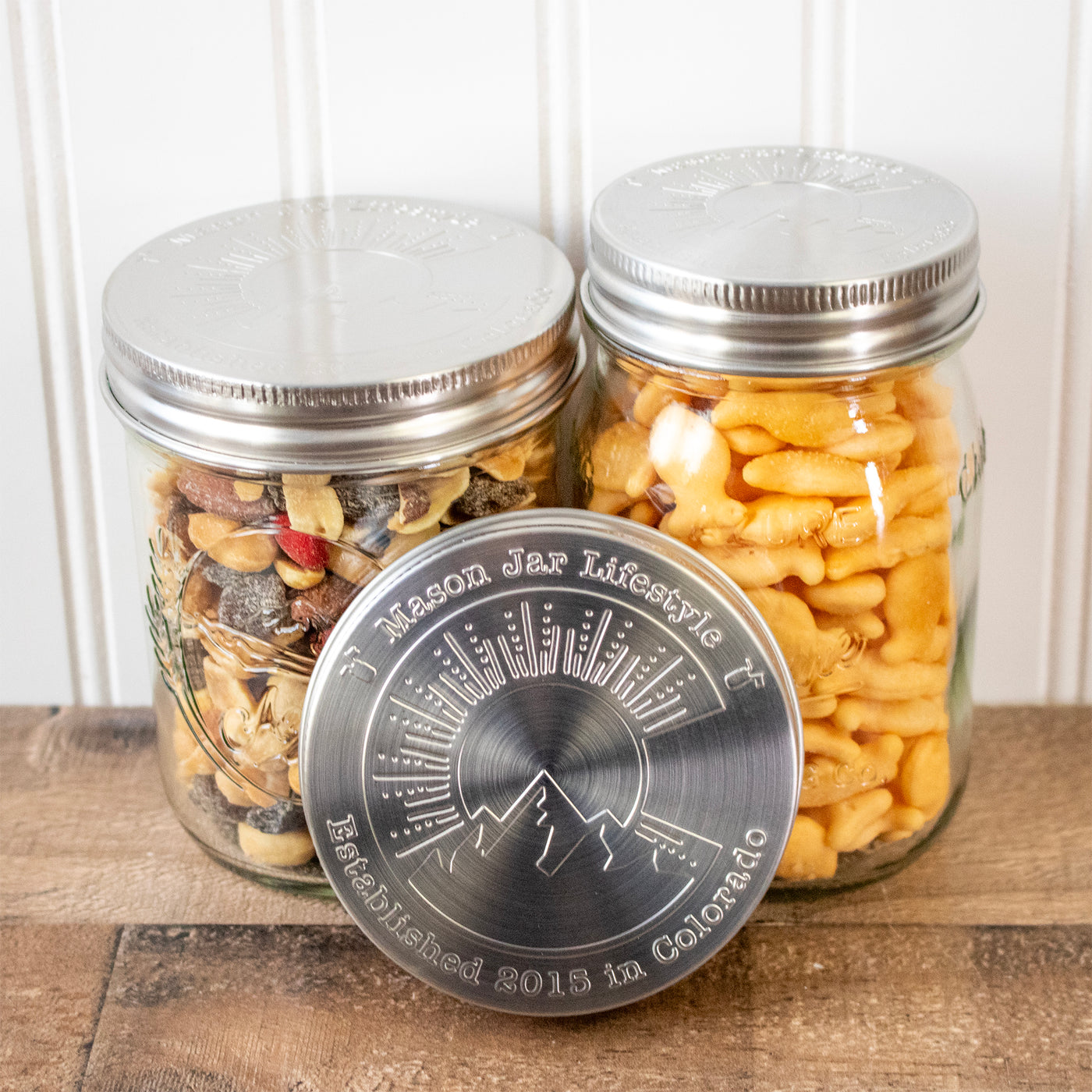 A regular mouth and wide mouth Mason jar with the Colorado storage lid on top, with snacks stored inside, and another lid propped up so you can see the design.