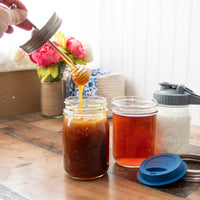 A hand pulling the dipper up from a regular mouth Mason jars full of honey, surrounded by other Mason jar accessories: a silicone drinking lid, and pour and store pitcher lid.