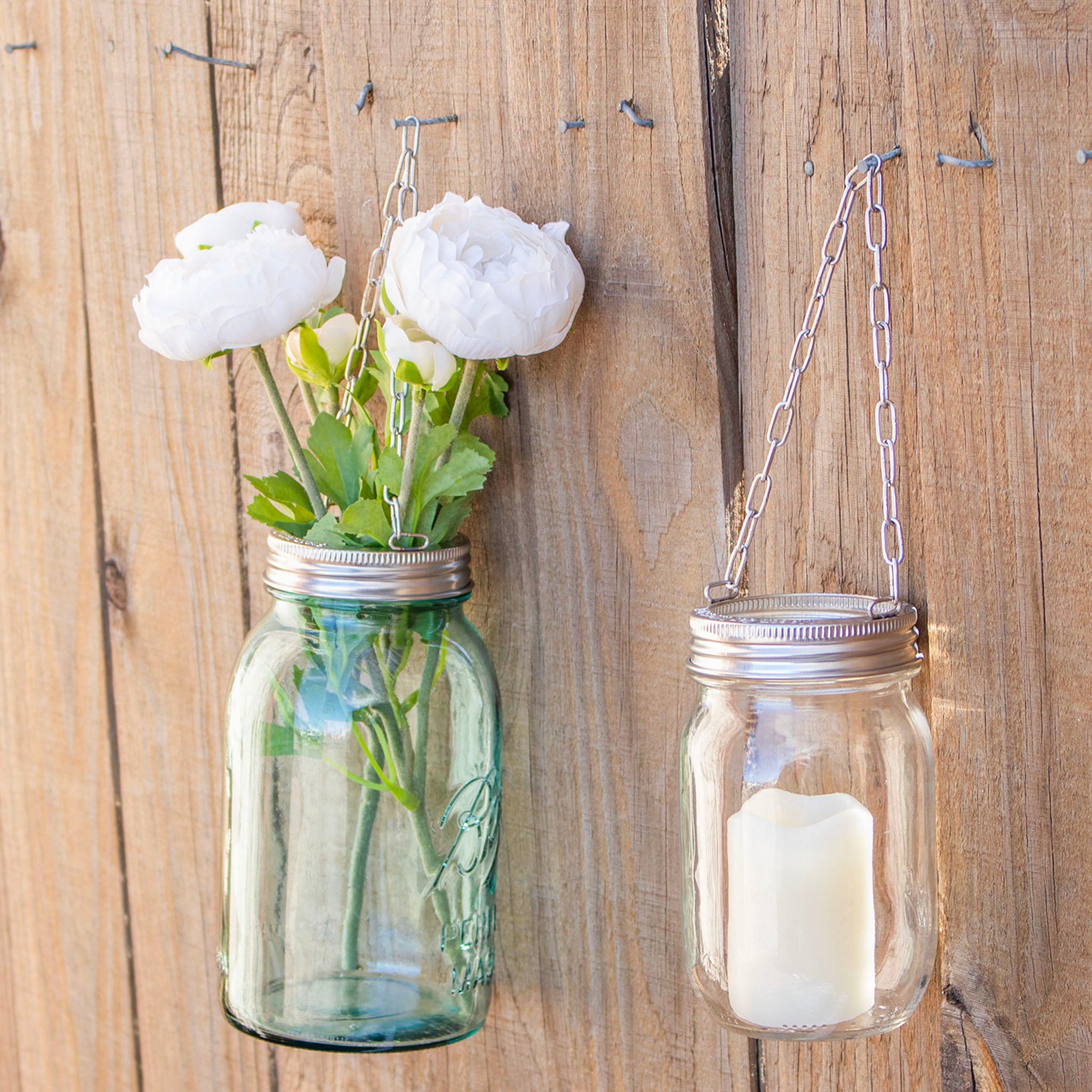 The band with chain on a Mason jar hung on a fence outside by a nail, with flowers and candles in them.