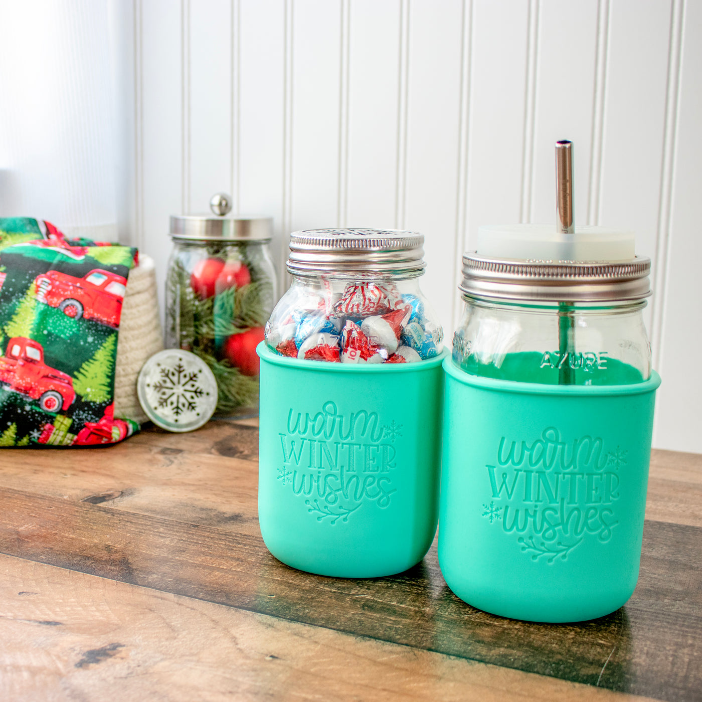 Two aquamarine sleeves with the Warm Winter Wishes design laser engraved into it, on a regular mouth and a wide mouth 32oz quart Mason jar. One is shown with candies inside and a decorative lid, the other is show with a silicone drinking lid and stainless steel straw. Behind is wintery decor in Christmas colors.