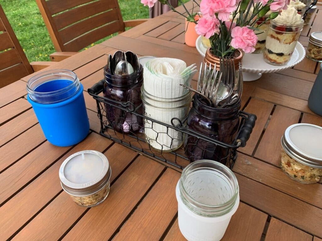 A backyard BBQ tablescape set up for easy outdoor entertaining with silverware and napkins in Mason jars held in a chicken wire caddy, individual portions of orzo pasta salad, flowers arranged with a frog lid, and half pint Mason jars in silicone sleeves.