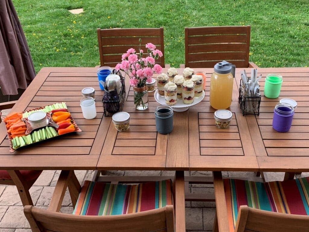 A backyard BBQ tablescape set up for easy outdoor entertaining with silverware, straws, and napkins in Mason jars held in a chicken wire caddy, individual portions of orzo pasta salad, flowers arranged with a frog lid, half pint Mason jars in silicone sleeves, homemade lemonade in a half gallon Mason jar with a pour and store lid with handle, and veggies with homemade ranch dip.