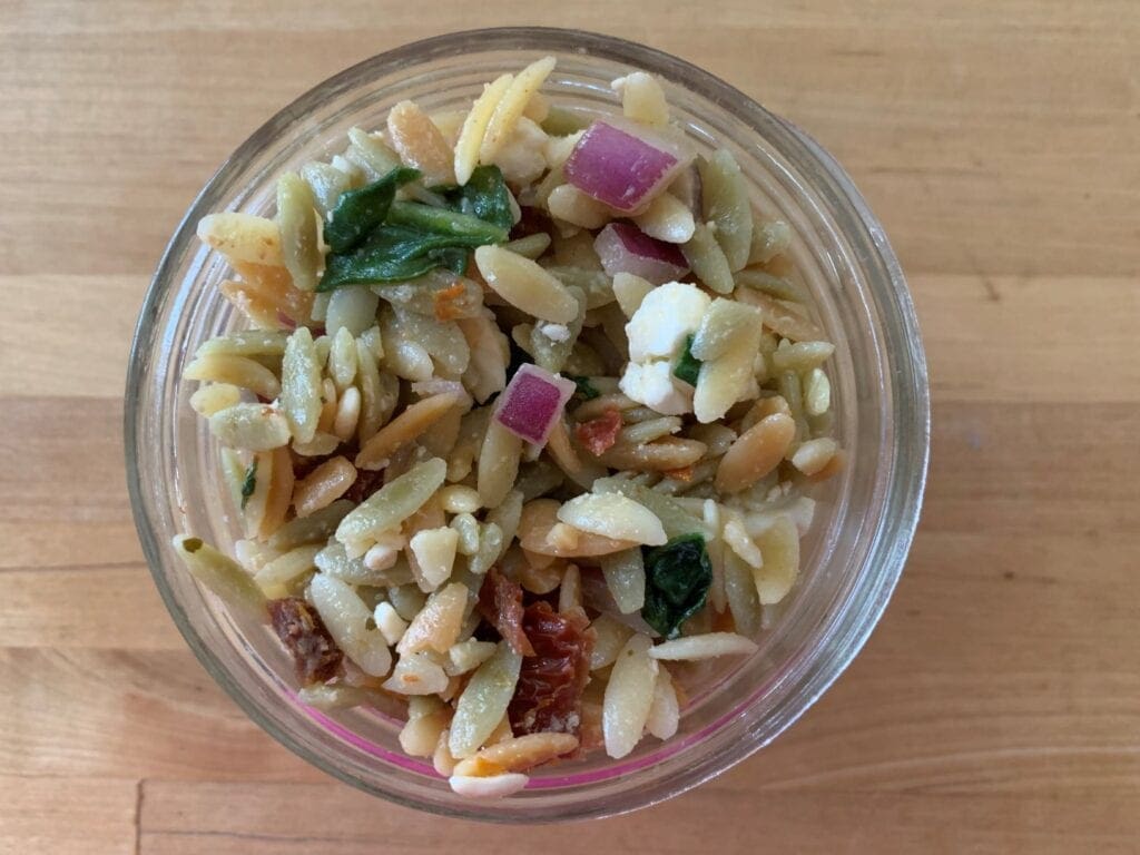 Orzo pasta salad that is easy to make with simple ingredients including tricolor orzo pasta, Italian dressing, spinach, feta cheese, red onion, sun-dried tomatoes in a wide mouth 8 oz half pint Mason jar.