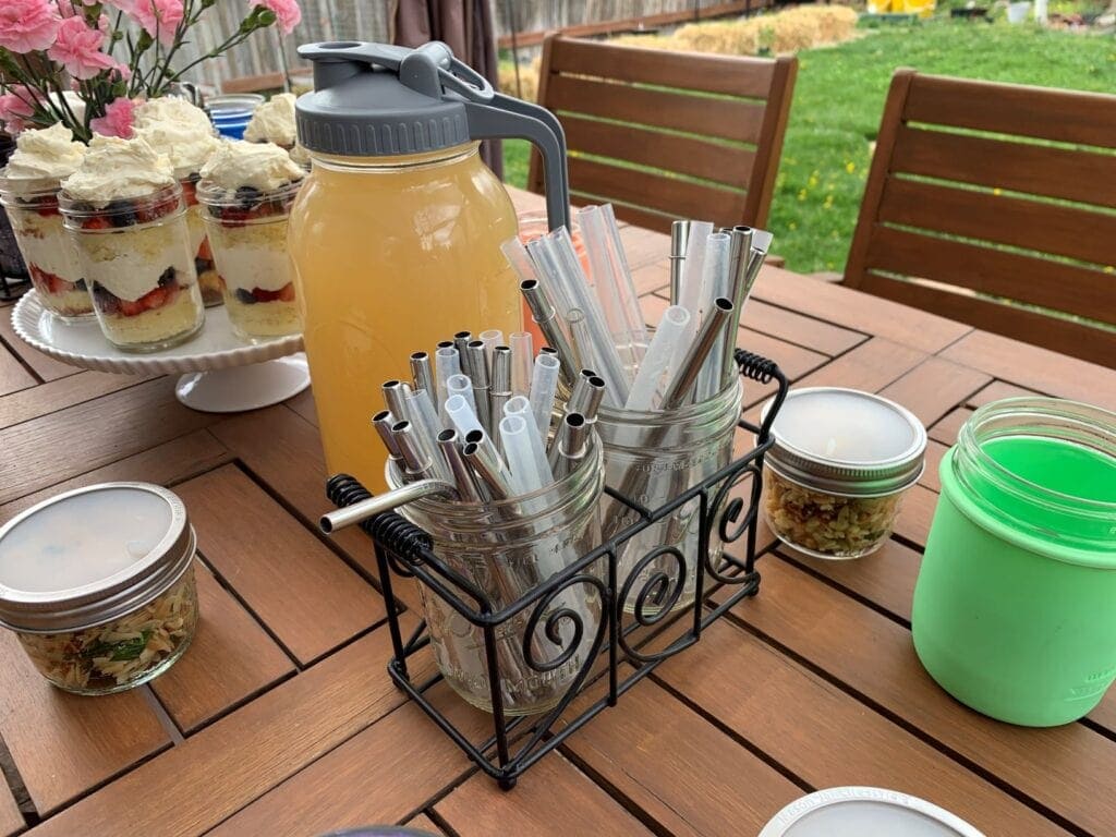 A backyard BBQ tablescape set up for easy outdoor entertaining with silverware, straws, and napkins in Mason jars held in a chicken wire caddy, individual portions of orzo pasta salad, flowers arranged with a frog lid, half pint Mason jars in silicone sleeves, homemade lemonade in a half gallon Mason jar with a pour and store lid with handle, and veggies with homemade ranch dip.